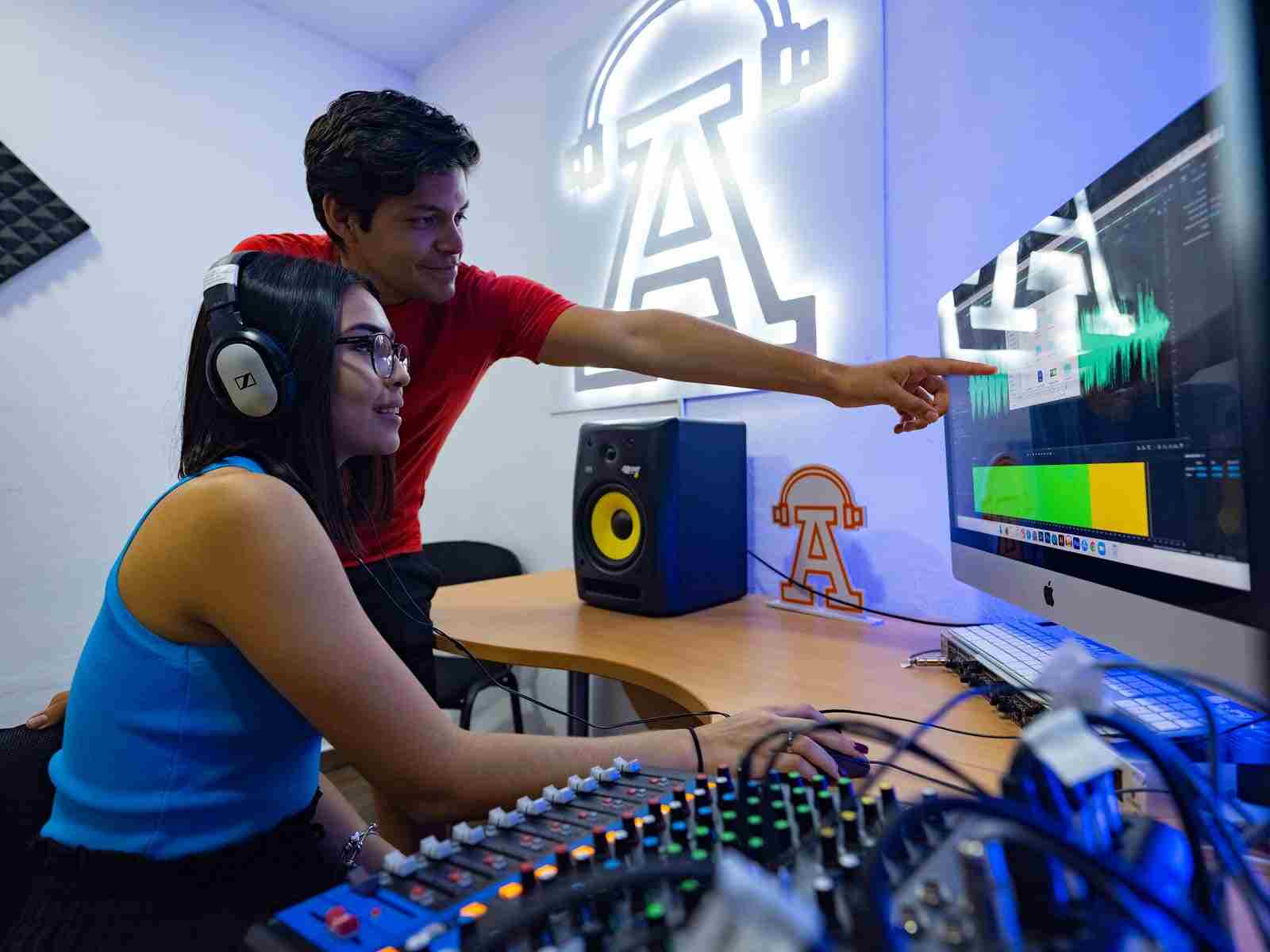 Estudiante con audífonos edita audio en una computadora mientras otro le señala la pantalla; ambos trabajan junto a una consola de mezcla en un estudio con logotipo iluminado.